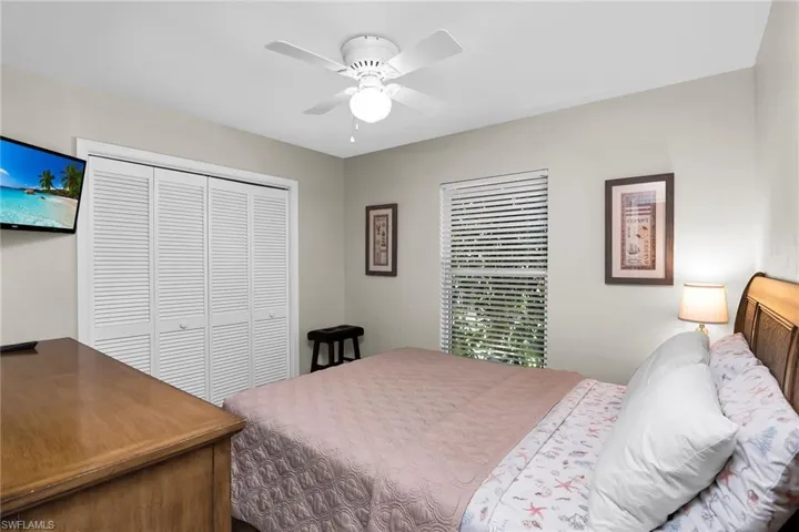 Louvered bi-fold closet doors, ceiling fan with light fixture, window with horizontal blinds, and wood-finish dresser with TV
