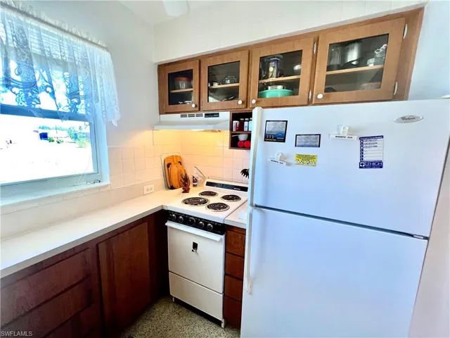 Kitchen featuring white appliances, light countertops, glass insert cabinets, under cabinet range hood, and backsplash