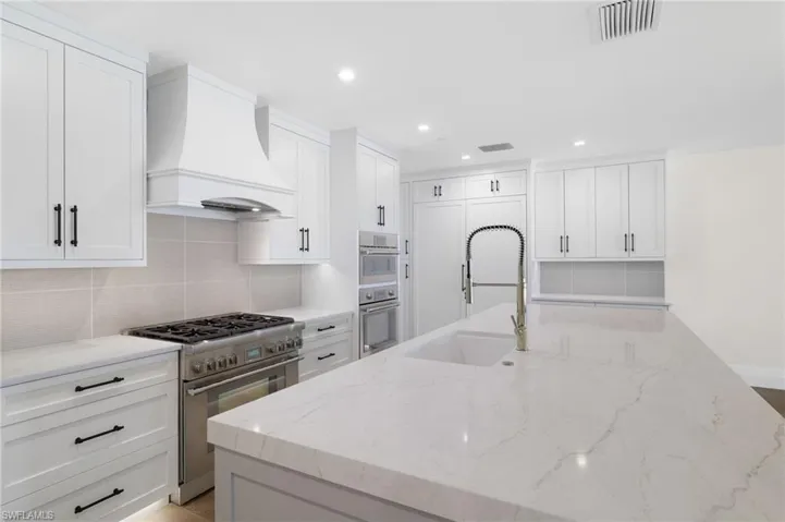 White cabinetry with black hardware, stainless steel range with hood, integrated wall oven and microwave, light-toned backsplash, and a kitchen island with an integrated sink and gooseneck faucet