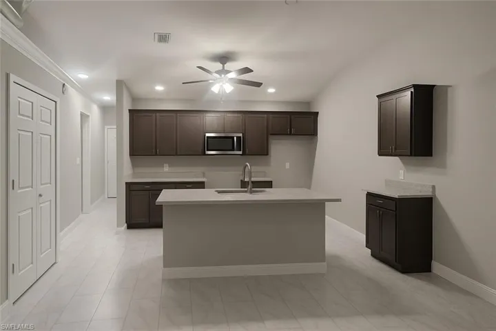 Kitchen featuring dark brown cabinetry, stainless steel microwave, a ceiling fan, light stone countertops, and a kitchen island with sink