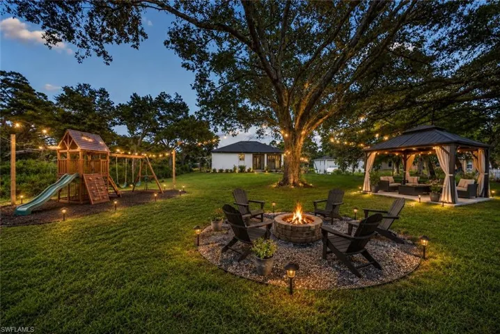Expansive yard featuring a stone fire pit, a gazebo, a play structure, and string lighting, all under mature trees