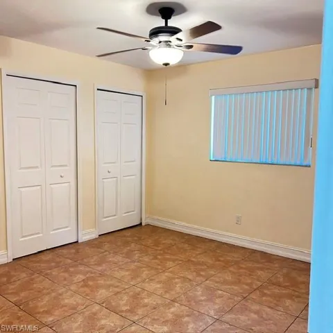 Bedroom featuring ceramic tile flooring, a window with vertical blinds, two bi-fold closet doors, and a ceiling fan with integrated lighting