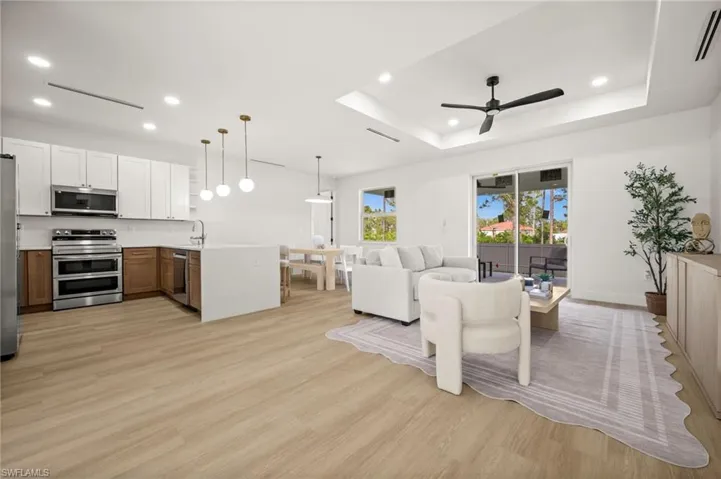This open-concept living space features light-toned wood style flooring, a kitchen with white upper and wood-tone lower cabinetry, stainless steel appliances, and pendant lighting