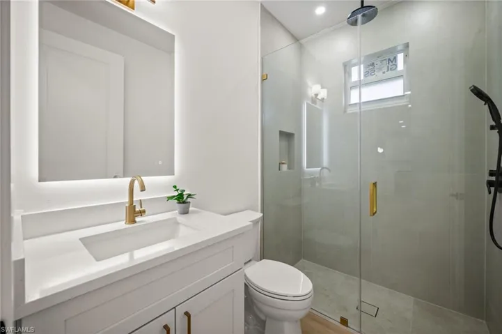 The bathroom features a modern vanity with a white countertop and gold-toned faucet, an illuminated mirror, and a spacious glass-enclosed shower with a black shower system