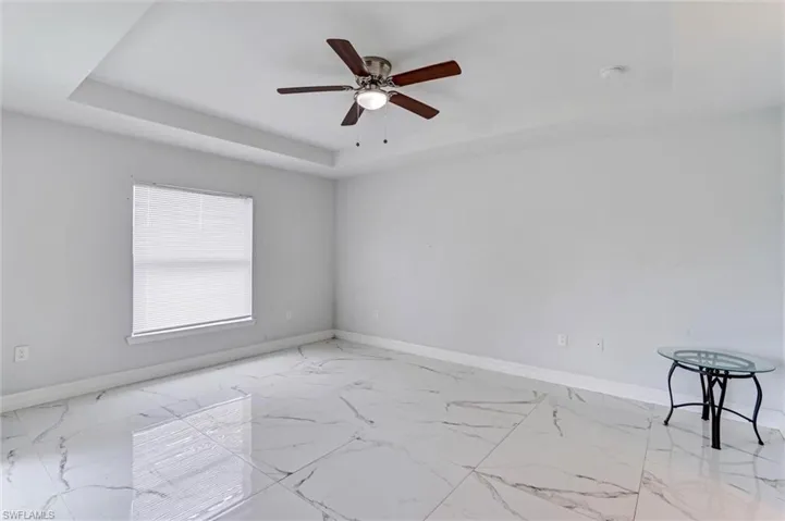Spacious room featuring large format tile flooring with distinctive veining, a tray ceiling, and a ceiling fan with integrated lighting