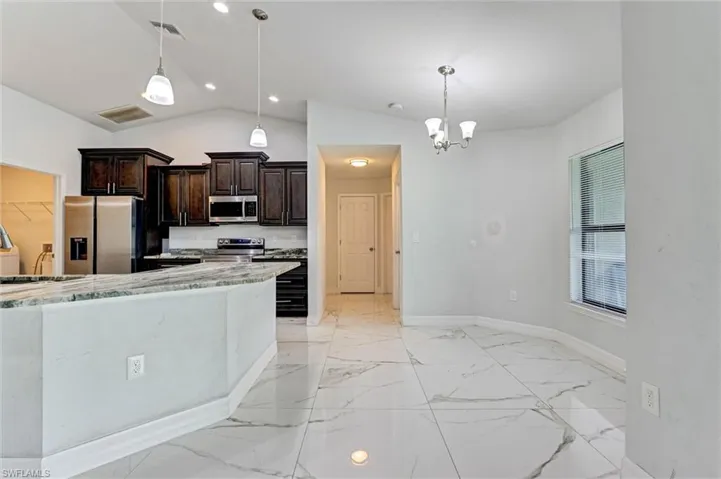 Open concept kitchen and dining area featuring vaulted ceilings, light-hued tile flooring, dark wood cabinetry, stainless steel appliances, and a breakfast bar with stone countertop