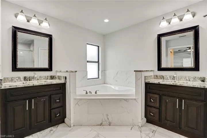 Bathroom featuring a corner soaking tub, dual dark wood-finish vanities with light stone countertops, and white wall and floor tile