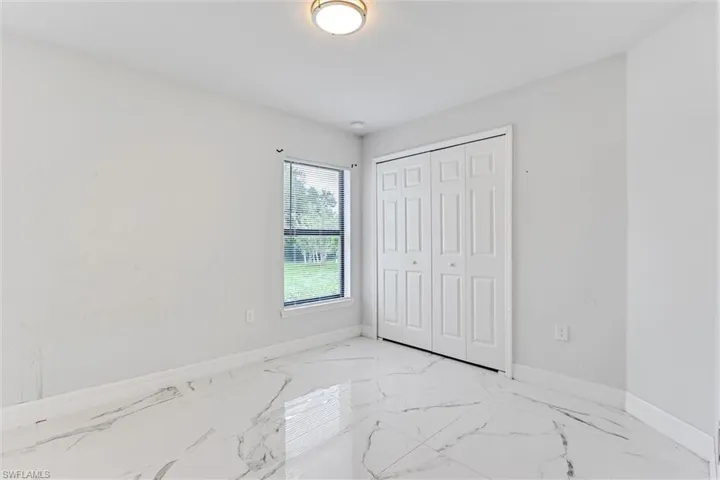 Spacious room featuring white marble-look flooring, large double-door closet, and a window with outdoor tree scenery