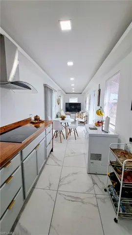 Kitchen featuring black electric cooktop, wall chimney exhaust hood, gray cabinetry, light marble finish flooring, and open floor plan
