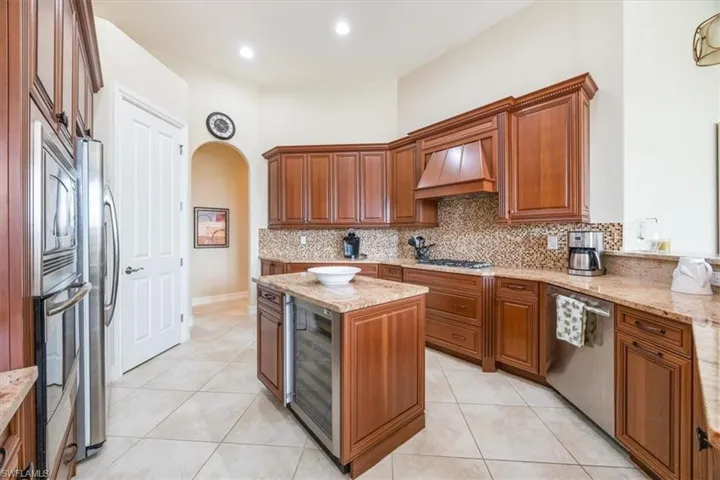 Kitchen with brown cabinetry, arched walkways, premium range hood, appliances with stainless steel finishes, and wine cooler