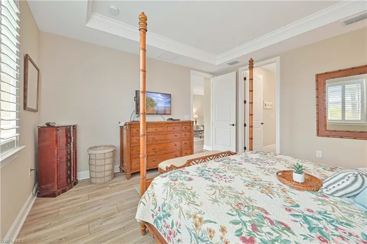 Bedroom featuring light wood finished floors, crown molding, a raised ceiling, baseboards, and ensuite bathroom