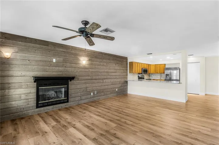 Wood-finish flooring throughout, a fireplace with a black mantel, a wood-paneled accent wall, a ceiling fan, and a kitchen pass-through with stone countertops