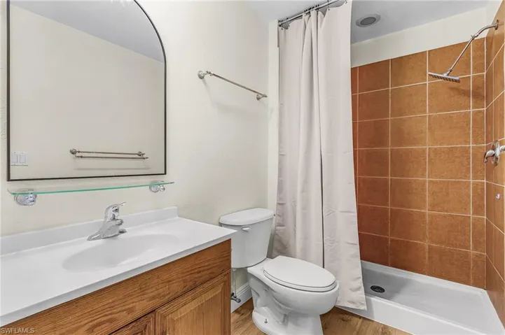 Vanity with a white countertop and wood-finish cabinetry, arched mirror, glass shelf, and a shower with brown tile surround
