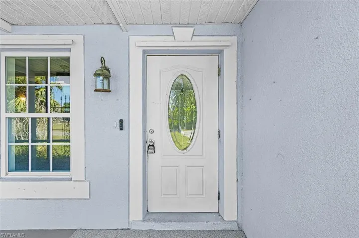 Textured exterior wall finish, white framed window with grilles, white entry door with oval glass insert, wall-mounted outdoor lantern, and a doorbell