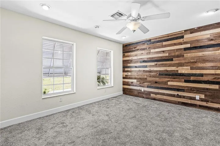 Accent wall with horizontal wood planks, two windows with blinds, recessed lighting, ceiling fan with light fixture, and gray carpet flooring