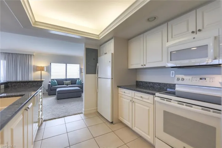 Kitchen with white cabinetry, a raised ceiling, dark stone counters, white appliances, and light tile patterned floors