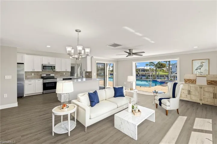 Open-concept living area featuring wood-finish flooring, a contemporary kitchen with white cabinetry and stainless steel appliances, and large sliding glass doors overlooking a pool and canal