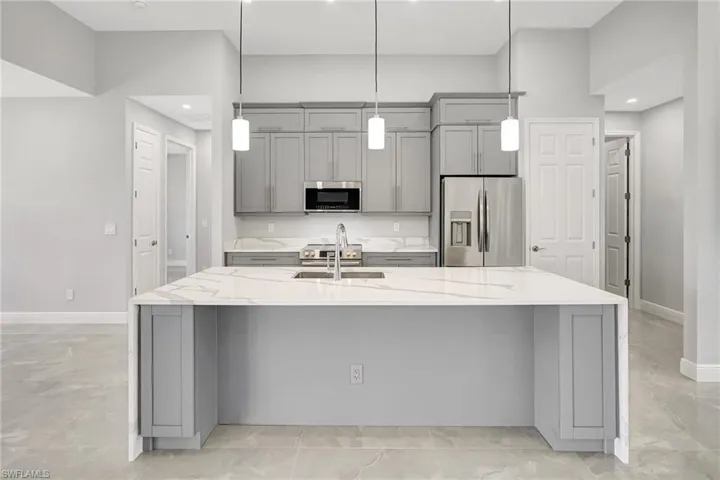 Kitchen with gray cabinets, light stone countertops, stainless steel appliances, an island with sink, and decorative light fixtures