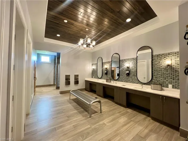 Full bath with double vanity, suspended lighting, light wood finished floors, decorative backsplash, and a wood tray ceiling