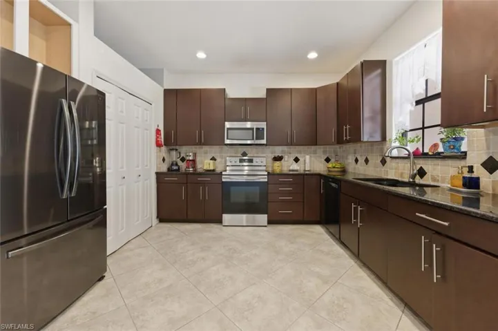 Kitchen featuring stainless steel appliances, dark wood finish cabinetry, dark stone countertops, light tile patterned flooring, and tasteful backsplash