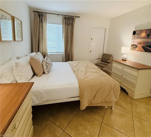 Bedroom featuring light-toned tile flooring, a window with blinds and patterned drapes, a white paneled door, and neutral-toned wall paint