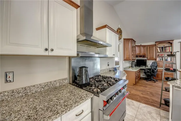 Kitchen with wall chimney range hood, lofted ceiling, high end range, light stone counters, and brown cabinets