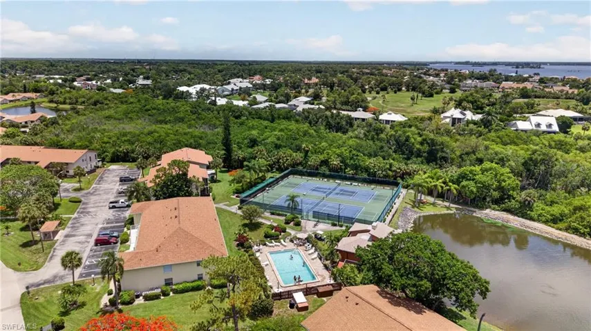 Aerial perspective highlighting a multi-building property featuring ample parking, a tennis court, a swimming pool, and a natural pond