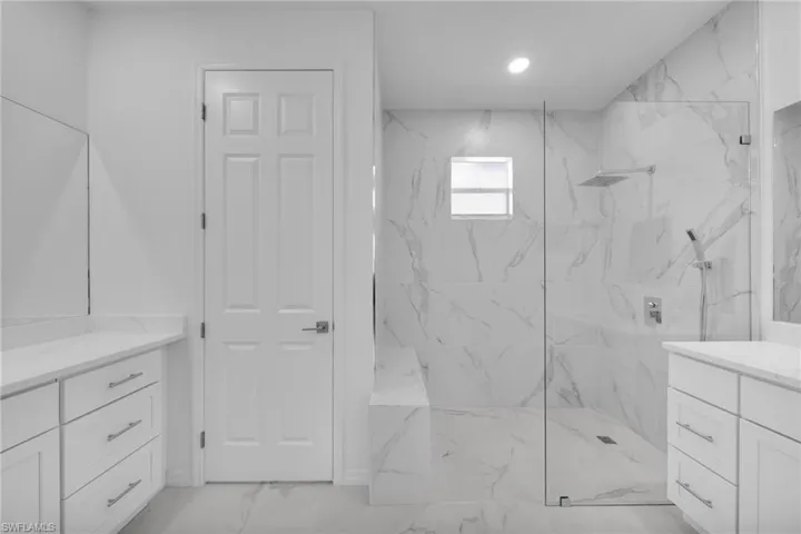 Spacious bathroom featuring a frameless glass shower enclosure with a rainfall showerhead, dual vanities with white cabinetry and light countertops, and large format marble-look tile flooring and shower surround