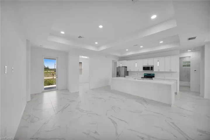 Open-concept living space featuring large format marble-look tile flooring, a spacious kitchen island with integrated sink, white cabinetry, stainless steel appliances, and tray ceilings with recessed lighting