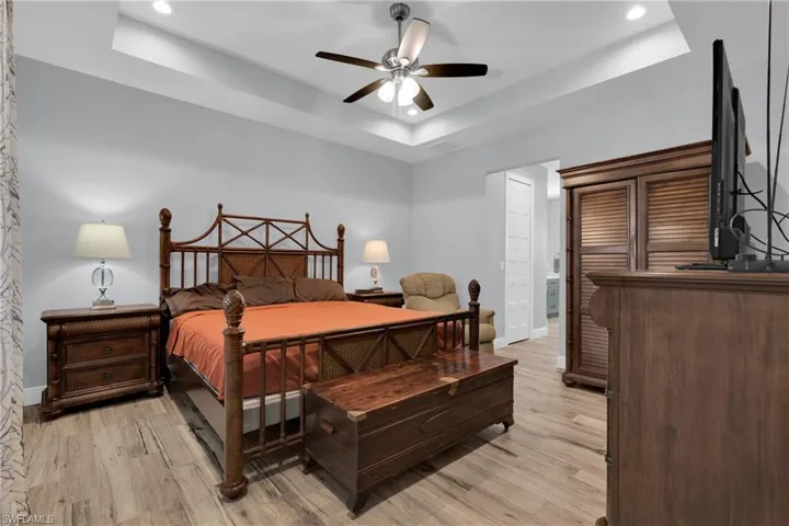 Bedroom with light wood-type flooring, a ceiling fan, a tray ceiling, ensuite bath, and recessed lighting