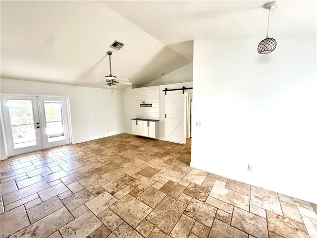 Unfurnished living room featuring a barn door, french doors, ceiling fan, stone tile flooring, and vaulted ceiling