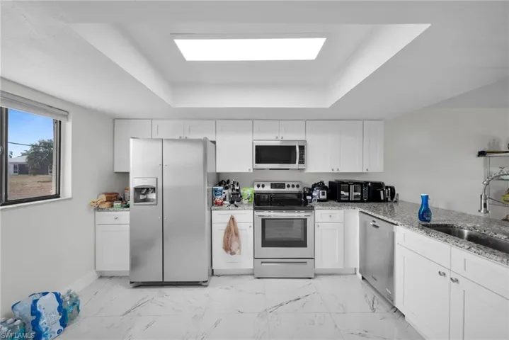 Kitchen featuring light stone countertops, stainless steel appliances, light marble finish floors, a raised ceiling, and white cabinetry