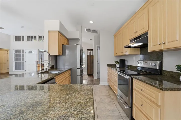 Kitchen with appliances with stainless steel finishes, light brown cabinetry, dark stone counters, under cabinet range hood, and vaulted ceiling