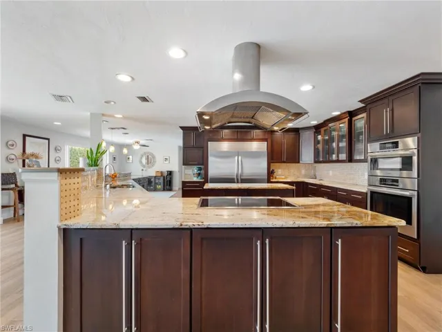 Kitchen featuring light wood finished floors, glass insert cabinets, dark wood finish cabinets, island exhaust hood, and light stone countertops