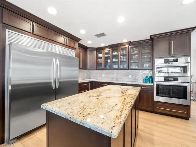 Kitchen featuring dark wood finish cabinets, stainless steel appliances, glass insert cabinets, light stone counters, and a center island