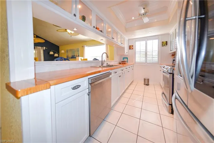 Galley kitchen featuring white cabinetry, stainless steel appliances, wood-finish countertops, tiled flooring, and a window with plantation shutters
