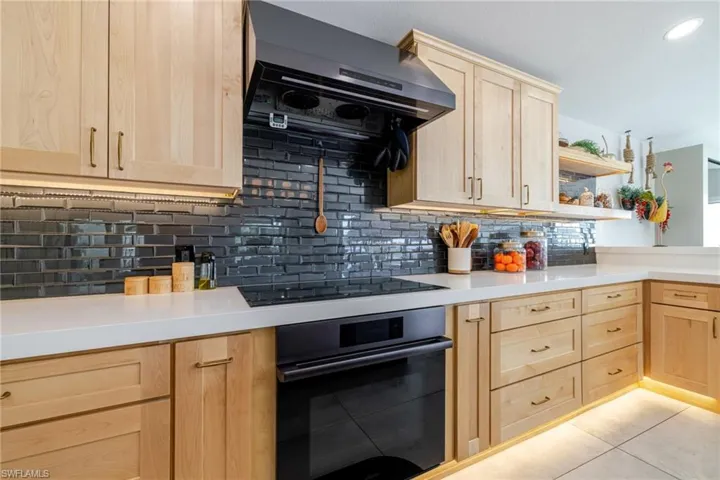 Kitchen with light wood finish cabinetry, black appliances, exhaust hood, open shelves, and tasteful backsplash