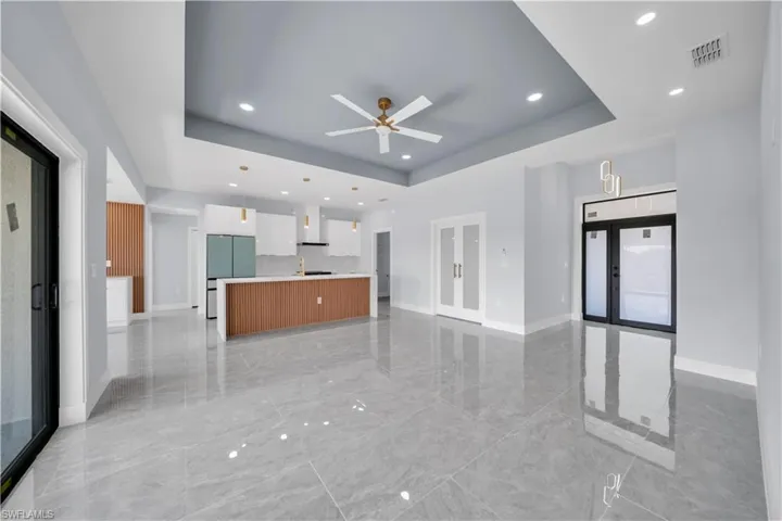Open-concept living space featuring large format gray tile flooring, a tray ceiling with recessed lighting and a ceiling fan, a kitchen island with wood-finish paneling, and white cabinetry