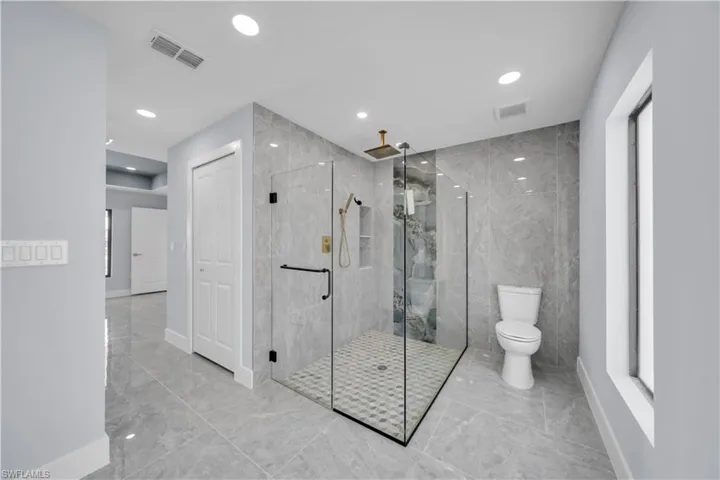Spacious bathroom featuring a glass-enclosed shower with gold-tone fixtures, floor-to-ceiling gray-toned tile, a separate toilet area, recessed lighting, and large format tile flooring