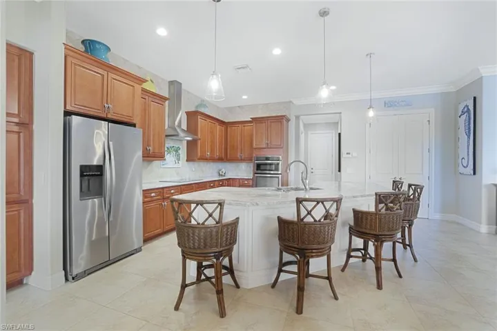 Kitchen with hanging light fixtures, stainless steel appliances, an island with sink, wall chimney exhaust hood, and light tile flooring