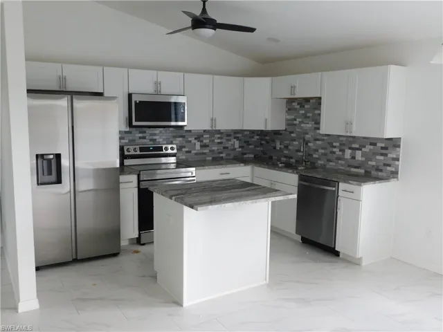 Kitchen featuring stainless steel appliances, light marble finish floors, lofted ceiling, dark stone counters, and white cabinetry