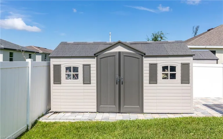 Exterior storage shed featuring a gray shingle-style roof, beige siding, double arched doors, and decorative windows with dark shutters