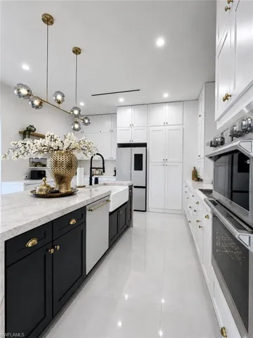 Modern kitchen featuring a large center island with a farmhouse sink, stone countertops, stainless steel appliances, white upper cabinetry, and contrasting black lower cabinetry