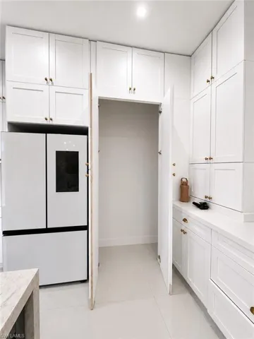 Bright kitchen area featuring extensive white shaker cabinetry with gold-tone hardware, a built-in pantry space, white large-format floor tiles, and a light-toned countertop