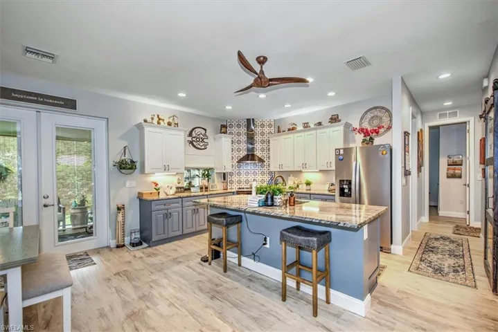 Kitchen featuring ceiling fan, a kitchen island with sink, light stone countertops, wall chimney exhaust hood, and light hardwood / wood-style floors