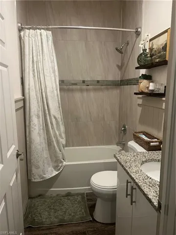 Full bathroom featuring a white porcelain tub, a tiled shower surround with accent band, a white vanity with granite-finish countertop, and wood-finish flooring