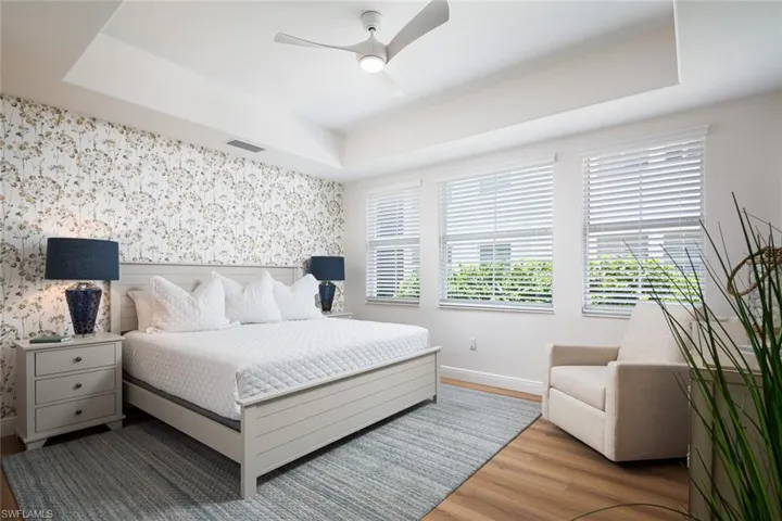 Bedroom with wood finished floors, wallpapered walls, a raised ceiling, multiple windows, and a ceiling fan