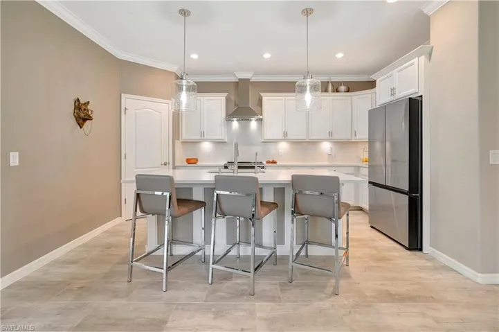 Kitchen featuring a kitchen bar, a center island with sink, stainless steel fridge, decorative light fixtures, and wall chimney exhaust hood