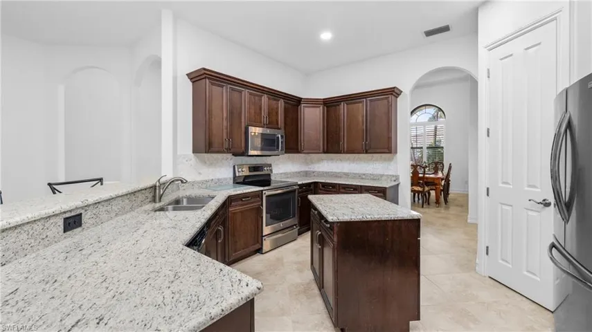 Kitchen featuring arched walkways, dark wood finish cabinetry, stainless steel appliances, a kitchen island, and light stone counters