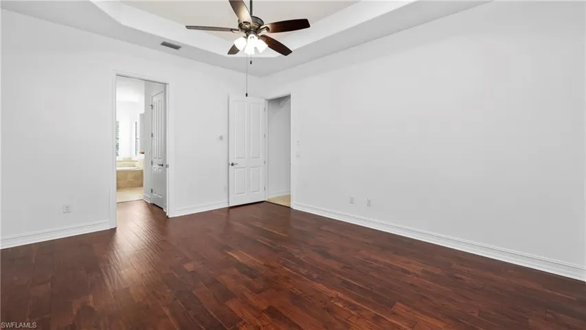Unfurnished bedroom with a raised ceiling, dark wood finished floors, ceiling fan, and ensuite bath